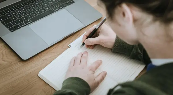 Nauczyciel pisze opinię o uczniu w zeszycie, obok leży laptop.