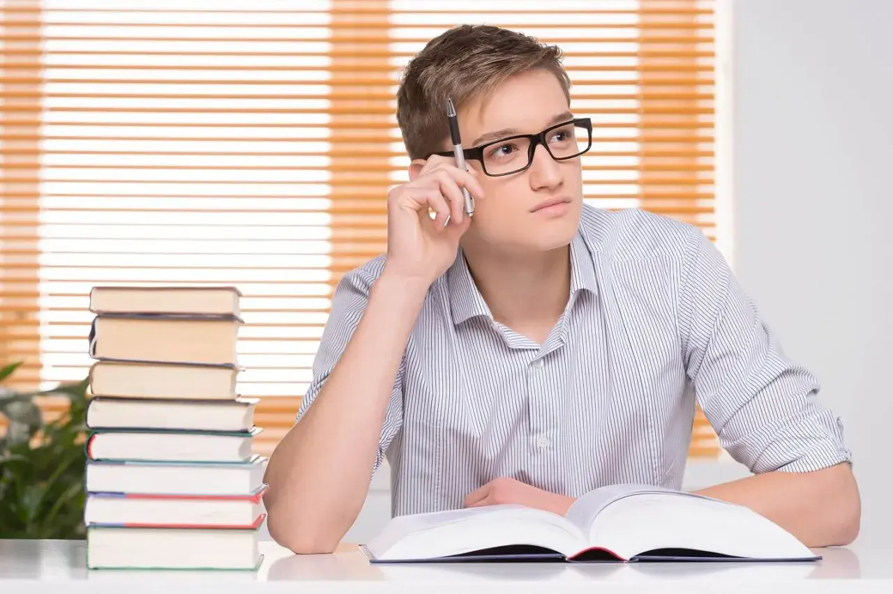 Młody mężczyzna w okularach z długopisem w dłoni zastanawia się nad tekstem w otwartej księdze. Obok stos książek, jakby przygotowywał się do egzaminu, który może wpłynąć na jego statut szkoły.