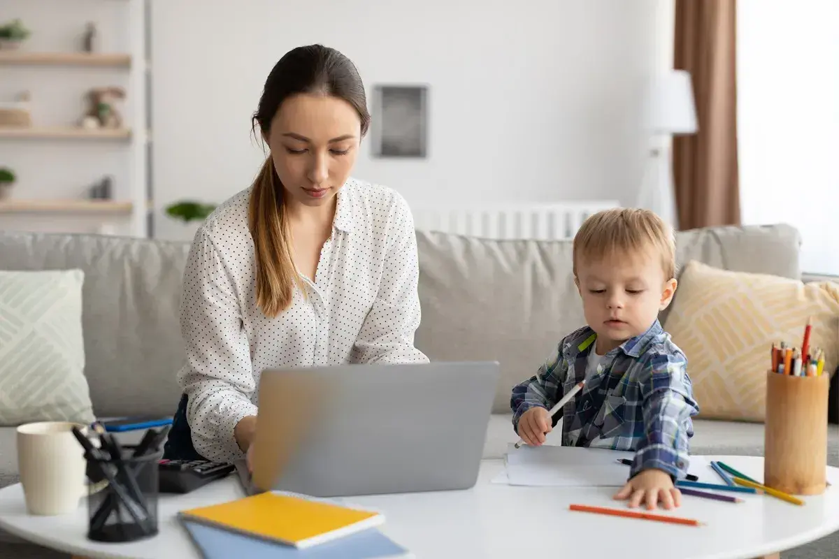 Kobieta pracuje na laptopie, podczas gdy jej syn rysuje obok. To codzienna opieka na dziecko i praca zdalna.