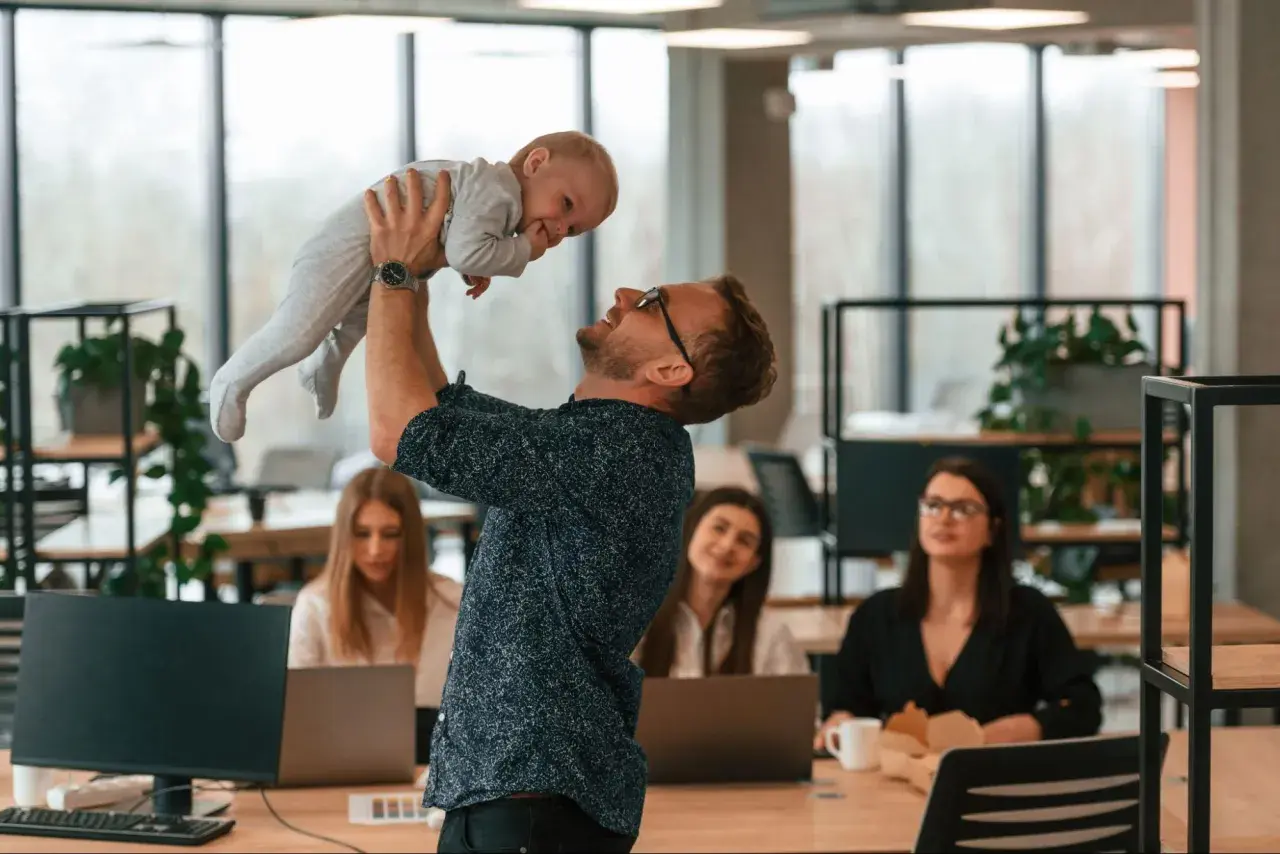 Szczęśliwy ojciec podrzuca dziecko w biurze, podczas gdy koleżanki pracują. Czy urlop wychowawczy wlicza się do stażu pracy?