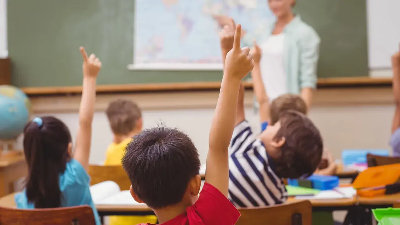 Uczniowie z podniesionymi rękami, gotowi do odpowiedzi. Nauczycielka ocenia ich zaangażowanie, co wpłynie na oceny z zachowania.