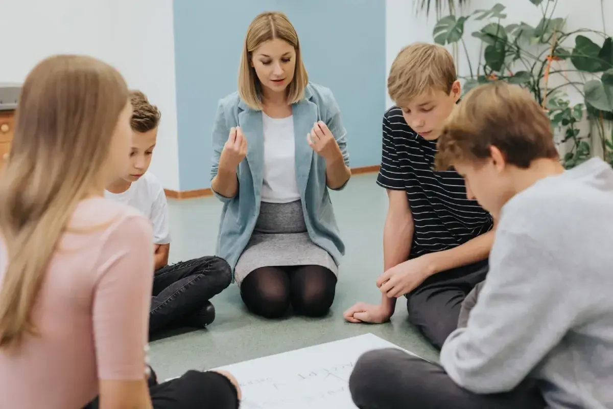 Nauczycielka z pasją tłumaczy coś grupie uczniów. Wszyscy siedzą na podłodze, skupieni na pracy.