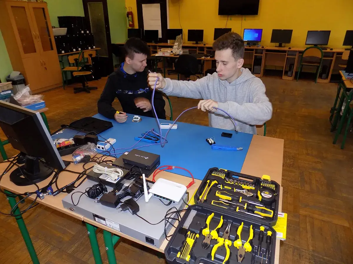 Dwóch uczniów ćwiczy technikę informatyczną, montując kable sieciowe. Na stole sprzęt i narzędzia.