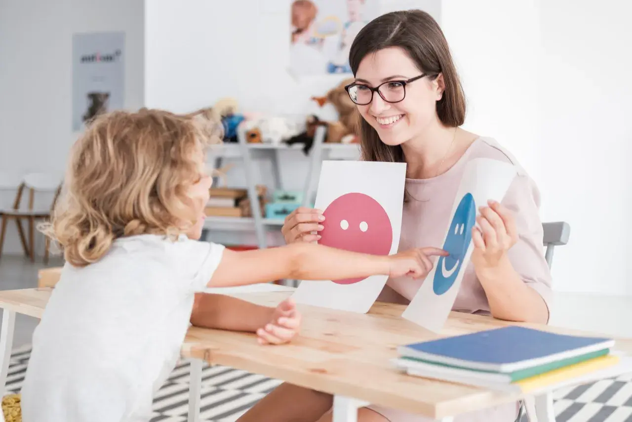 Uśmiechnięta psycholog z okularami pokazuje dziecku karty z buźkami. To część przygotowania pedagogicznego dla psychologów.