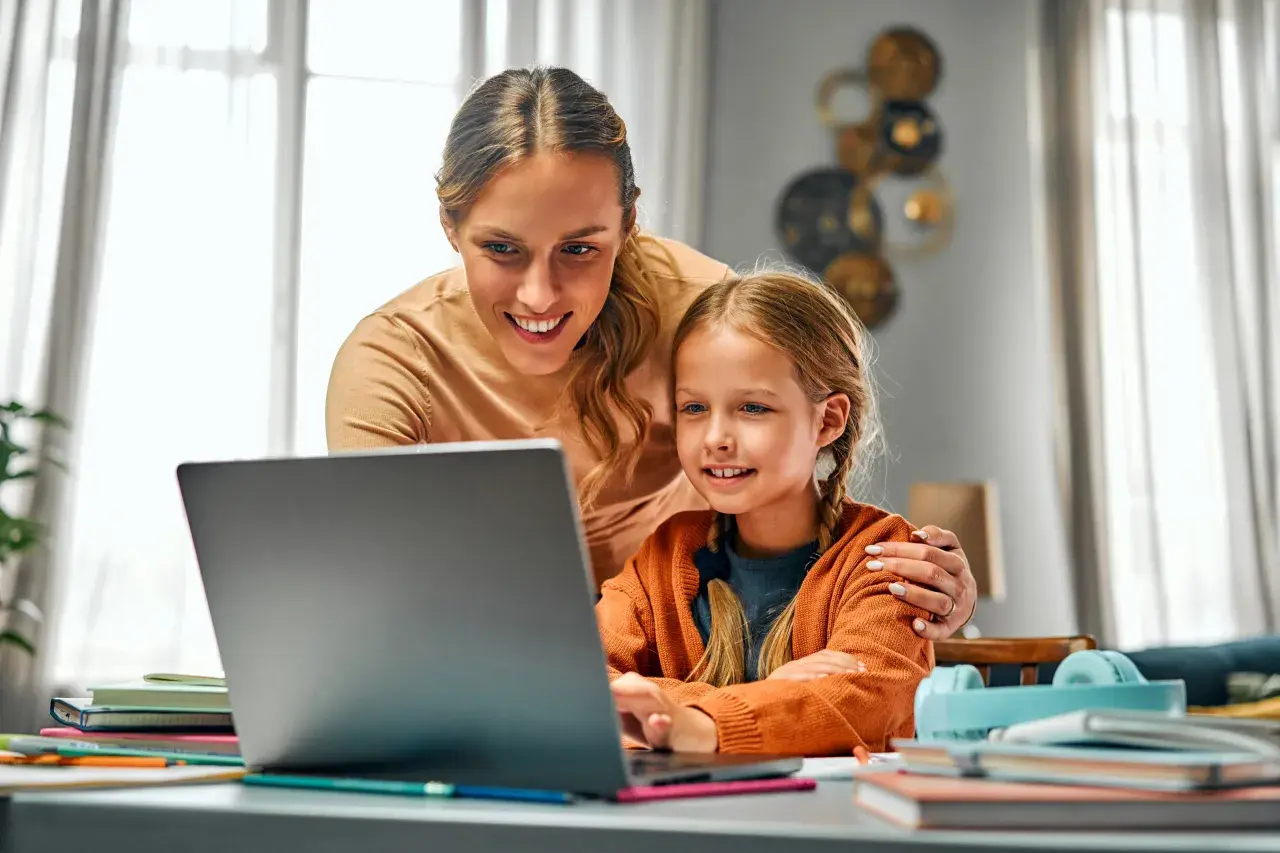 Uśmiechnięta mama pomaga córce w nauce przy laptopie. To przykład troskliwej opieki na dziecko 2 dni w tygodniu, gdy rodzice pracują.