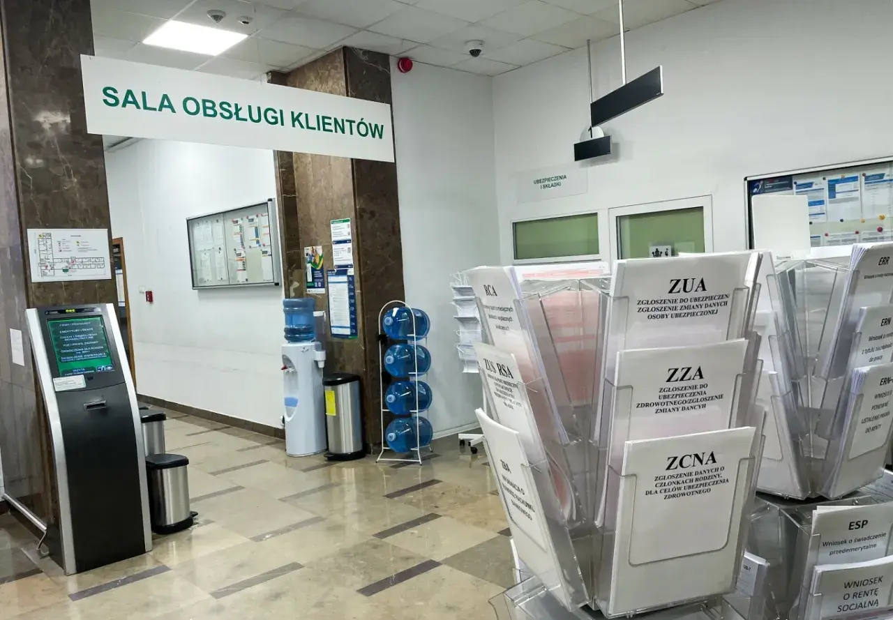 W sali obsługi klienta ZUS, obok dystrybutora wody, stoją stojaki z formularzami, m.in. ZUA, ZZA, ZCNA, RCA, ZUS RSA.