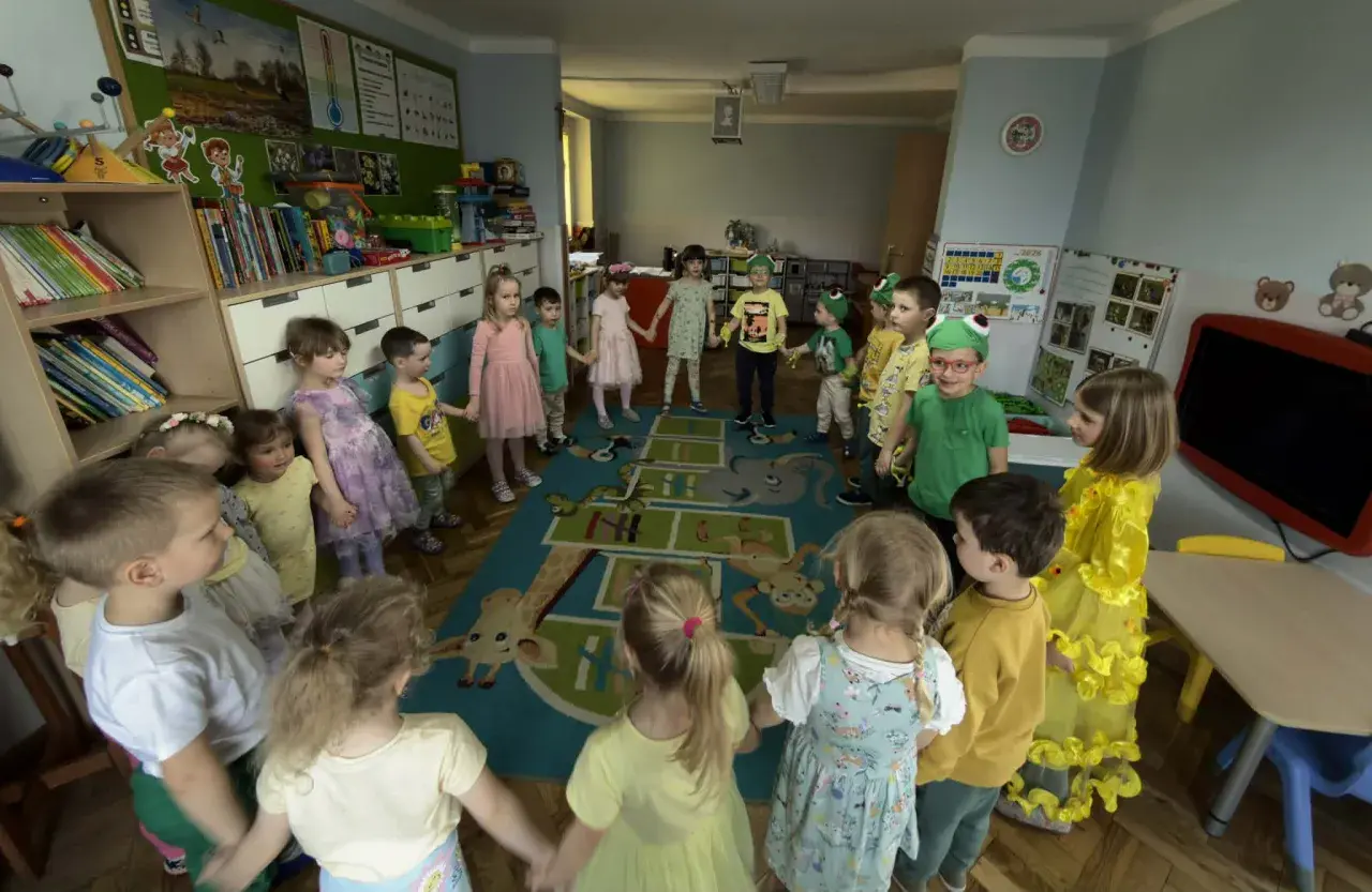 Dzieci trzymają się za ręce w kręgu na dywanie z zabawnymi zwierzątkami. To radosny dzień w **zespole szkolno przedszkolnym**.