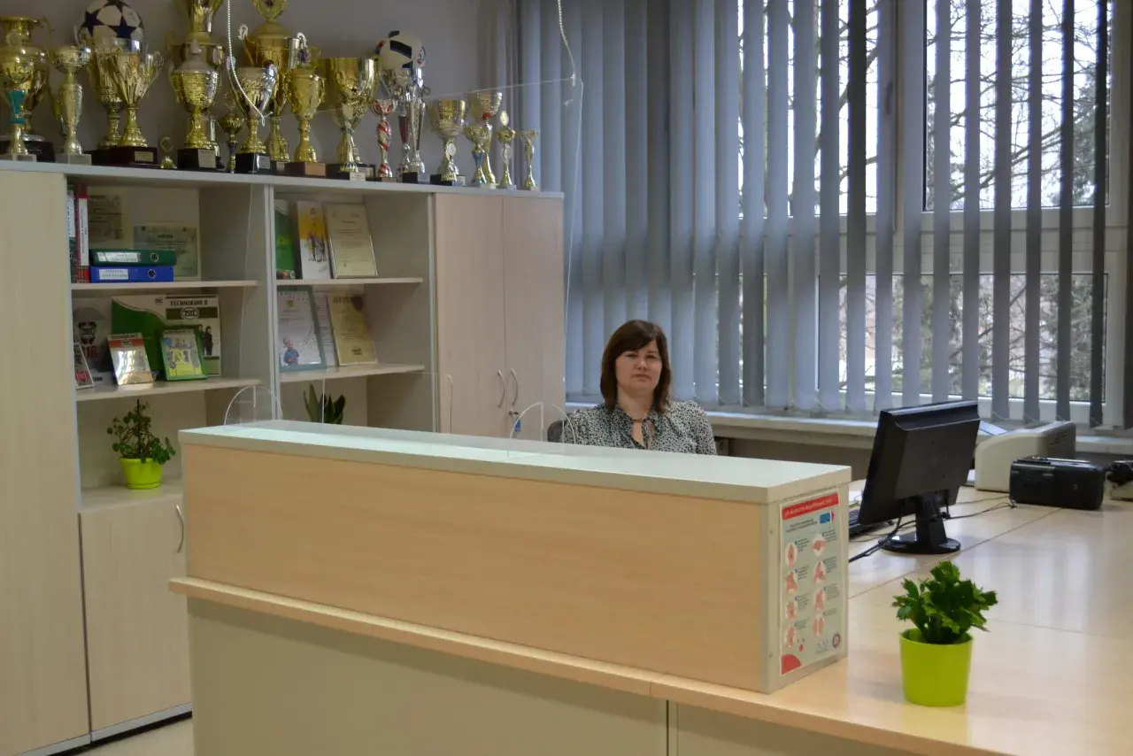Kobieta siedzi za ladą w sekretariacie, wśród licznych pucharów i dyplomów.