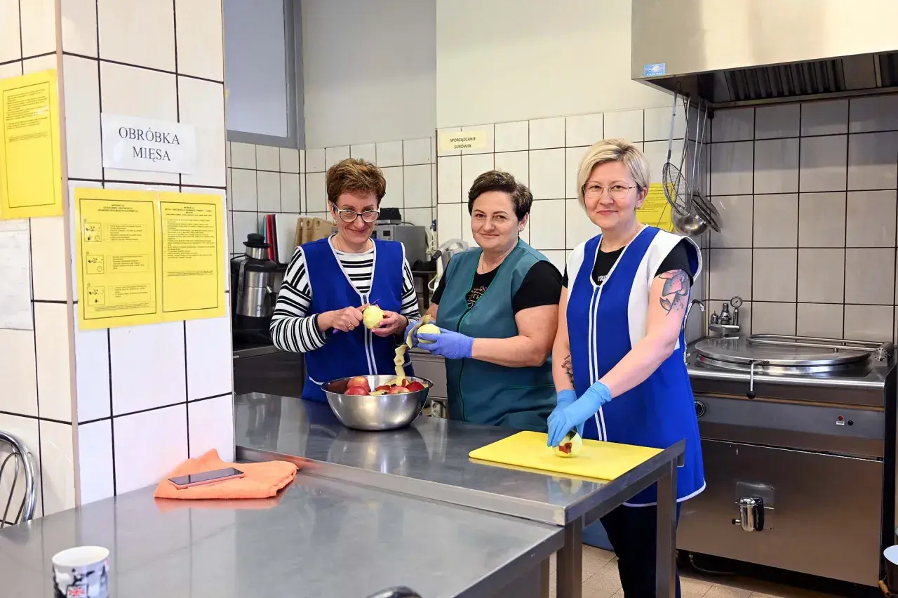 Trzy kobiety w fartuchach obierają jabłka w kuchni. Jedna z nich, intendentka, przygotowuje owoce do dalszej obróbki.