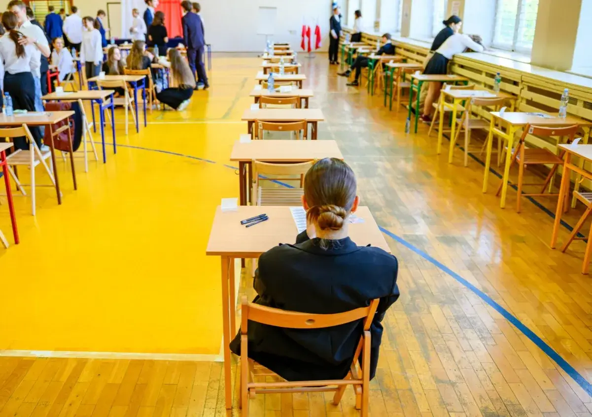 Uczniowie siedzą przy ławkach w sali gimnastycznej, przygotowując się do egzaminu dojrzałości.