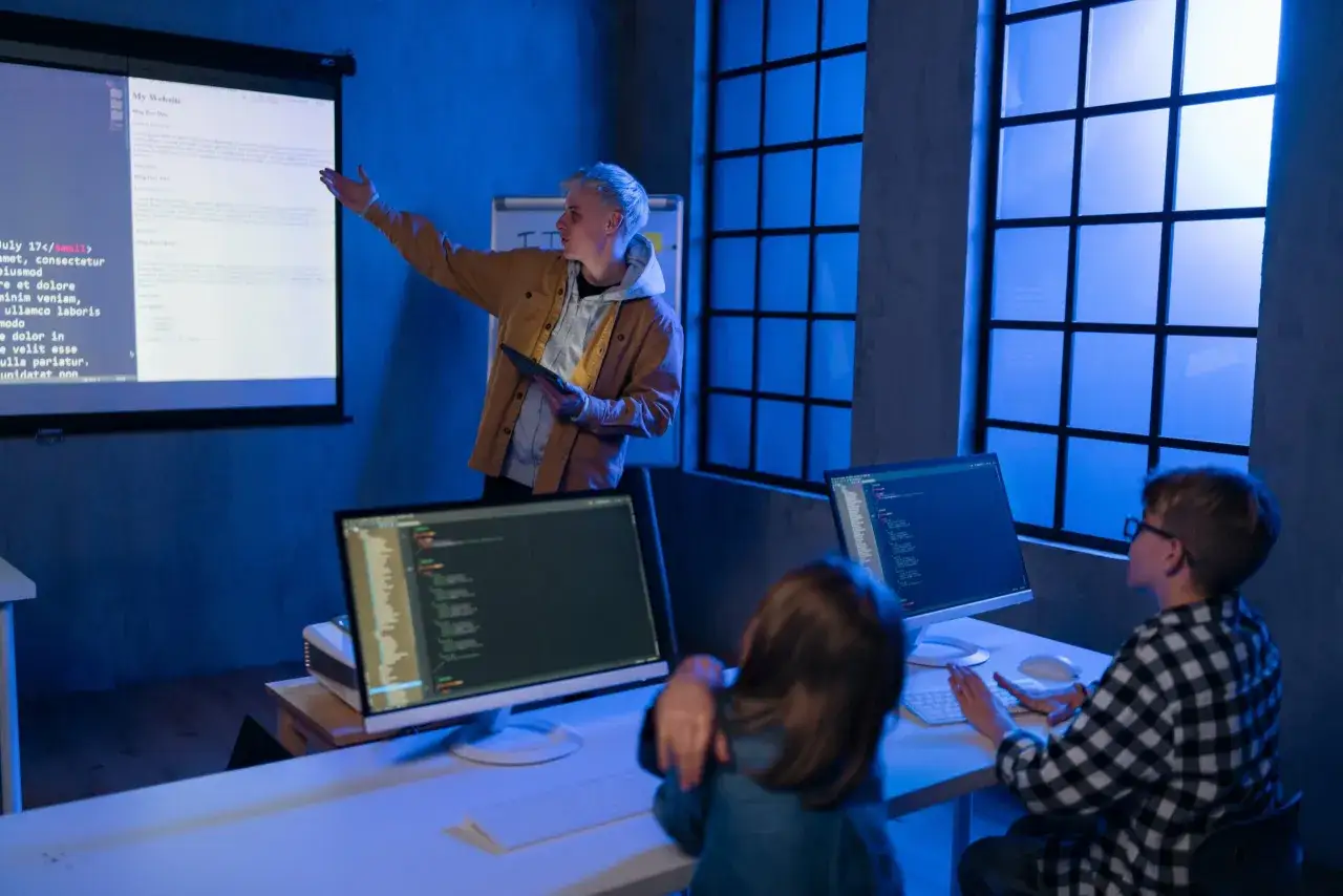 Młody technik programista prezentuje kod na ekranie, podczas gdy dwójka dzieci uczy się kodowania przy komputerach.
