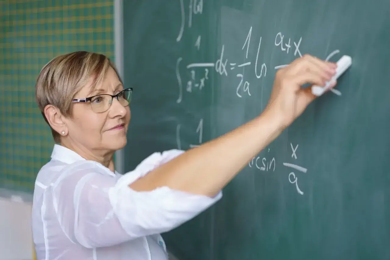 Nauczycielka z uśmiechem pisze kredą wzory matematyczne na tablicy. To obrazek symbolizujący nagrodę jubileuszową nauczycieli.