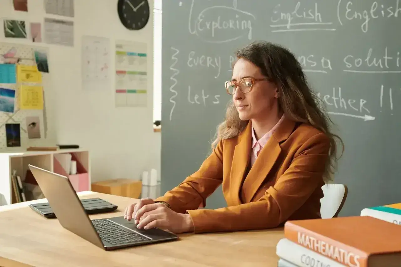 Nauczyciel początkujący w okularach pracuje przy laptopie w klasie. Na tablicy napisy po angielsku.