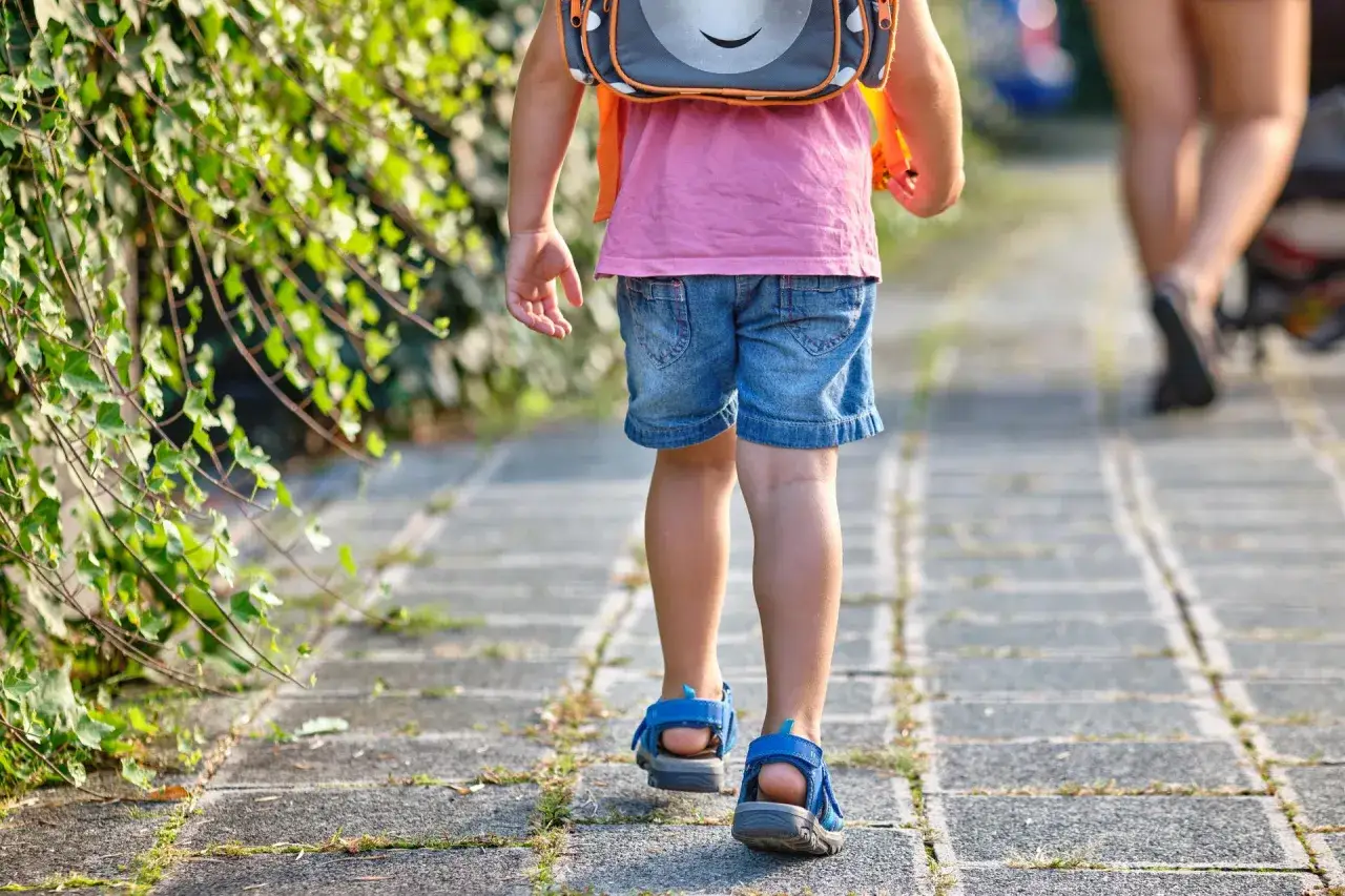 Małe dziecko w dżinsowych szortach i różowej koszulce, z plecakiem na plecach, idzie chodnikiem. To piękny dzień, kiedy dzieci idą do szkoły.
