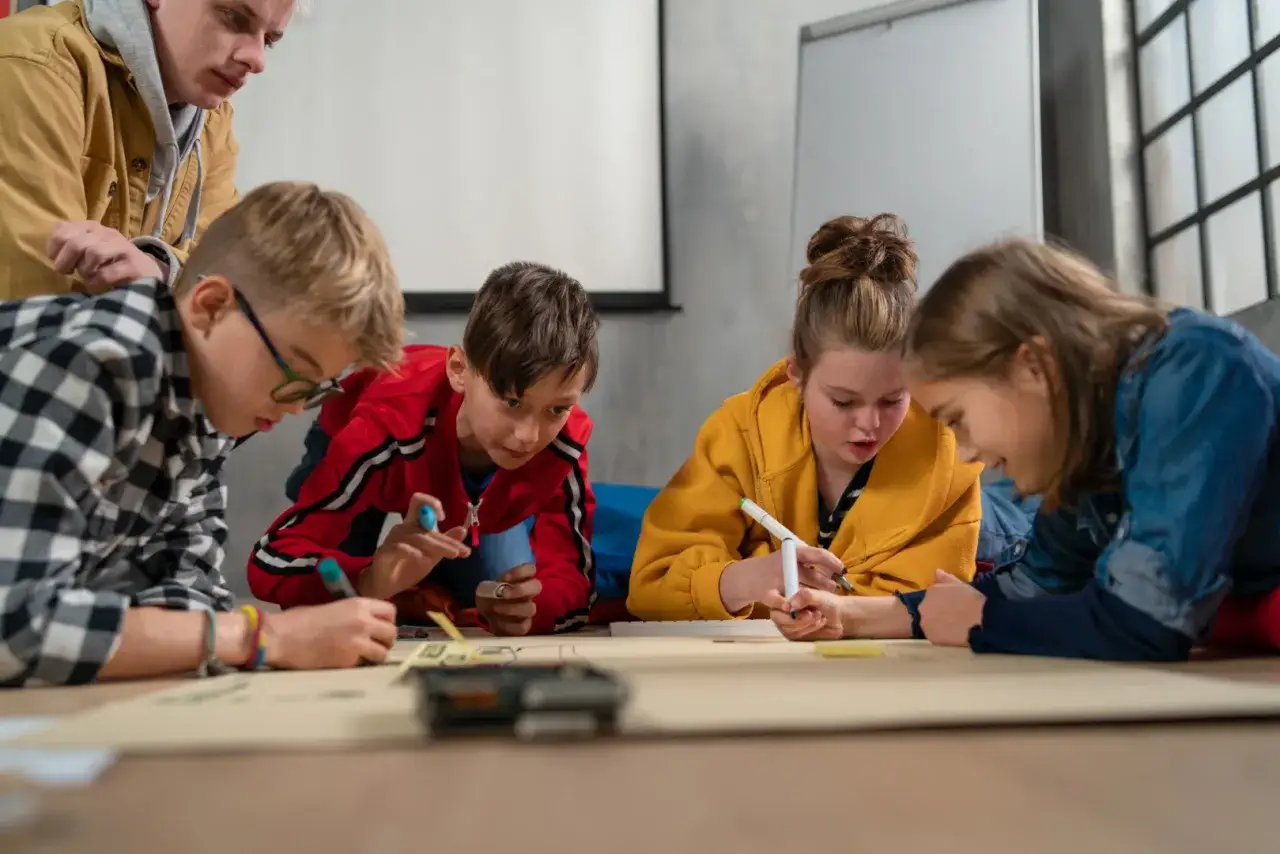 Dzieci z nauczycielem rysują na wielkim arkuszu papieru. Używają pisaków, by stworzyć projekt.