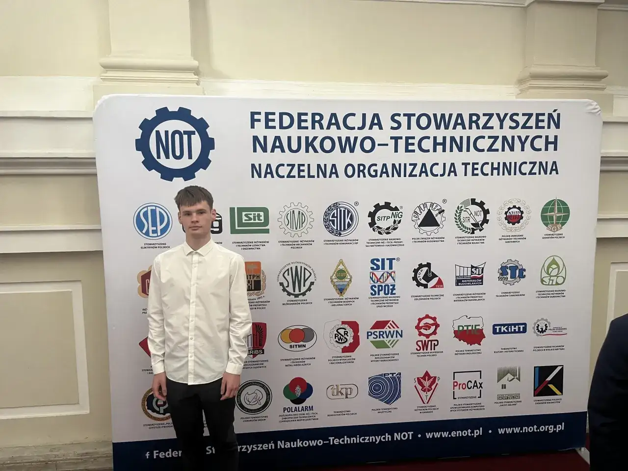 Młody mężczyzna na tle baneru Federacji Stowarzyszeń Naukowo-Technicznych NOT, symbolizującego rozwój w obszarze biznesu i zarządzania.