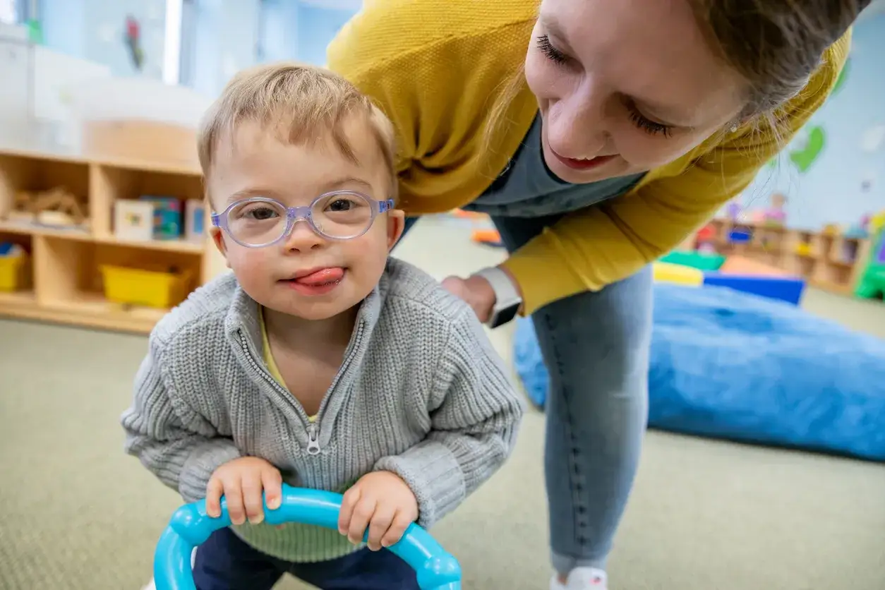 Uśmiechnięty chłopiec z zespołem Downa, w okularach, bawi się z oligofrenopedagogiem w sali terapeutycznej.