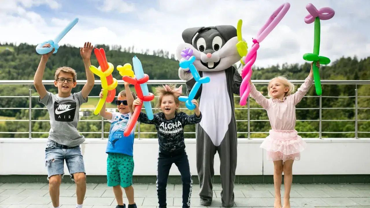 Radosne dzieci z balonami w kształcie zwierząt i mieczy świętują, kiedy kończy się rok szkolny. Wesoły królik w stroju maskotki dołącza do zabawy.