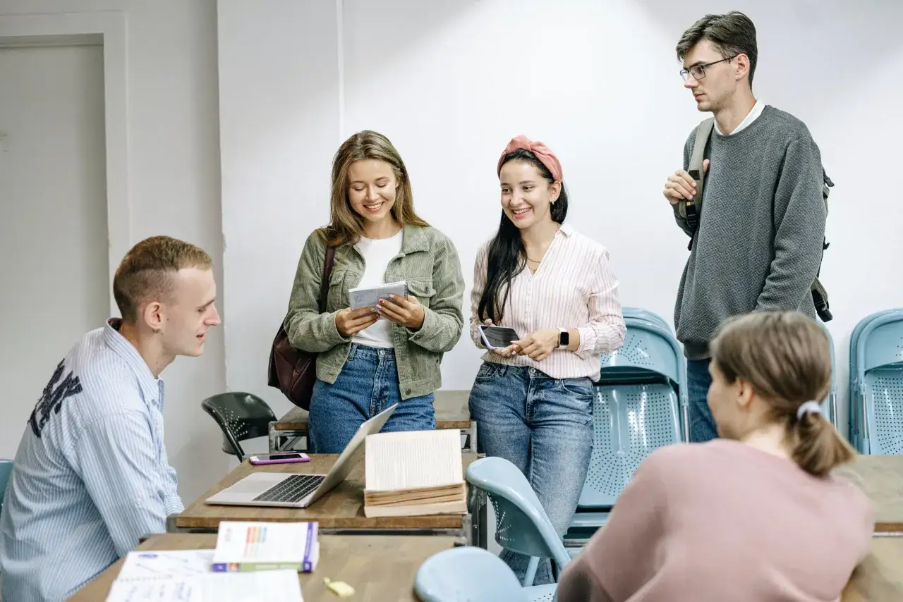 Studenci dyskutują przy stole z laptopem i książkami. W tle widać krzesła i innych uczestników spotkania, być może podczas zajęć z glottodydaktyki.