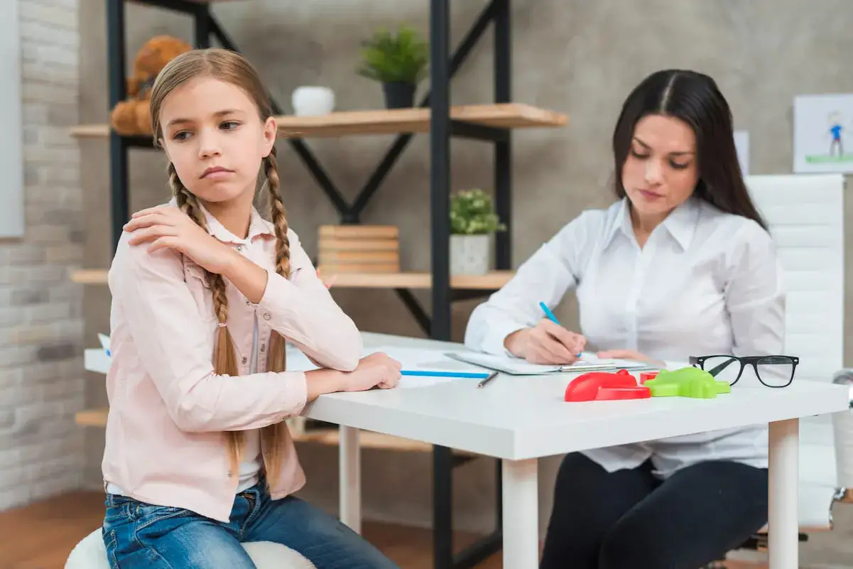 Dziewczynka z warkoczami siedzi przy biurku, zamyślona, podczas gdy psycholog robi notatki. Trwa diagnoza psychologiczna.