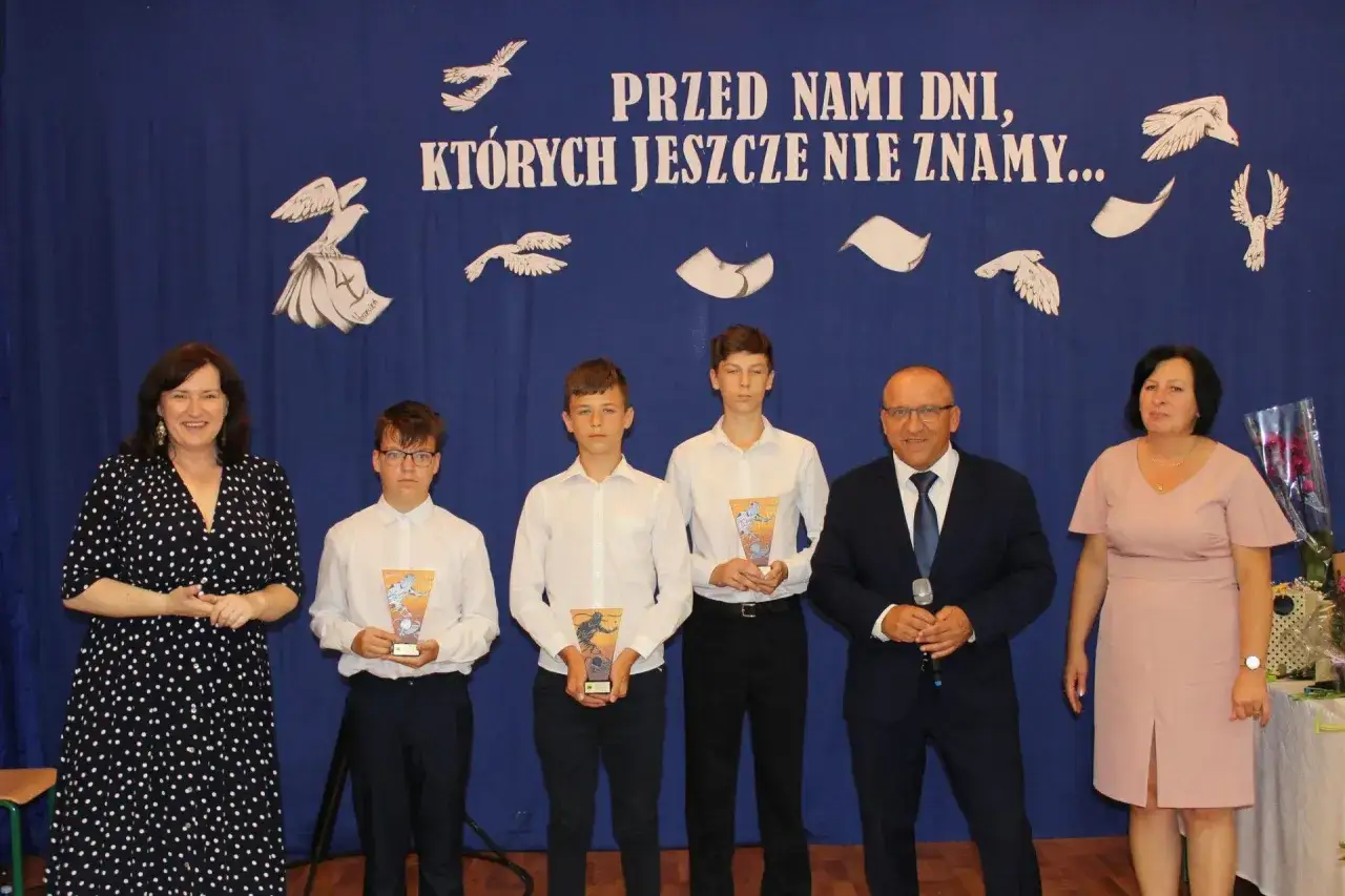 Uroczyste zakończenie roku szkolnego. Nauczyciele wręczają nagrody uczniom na tle niebieskiej kurtyny z napisem