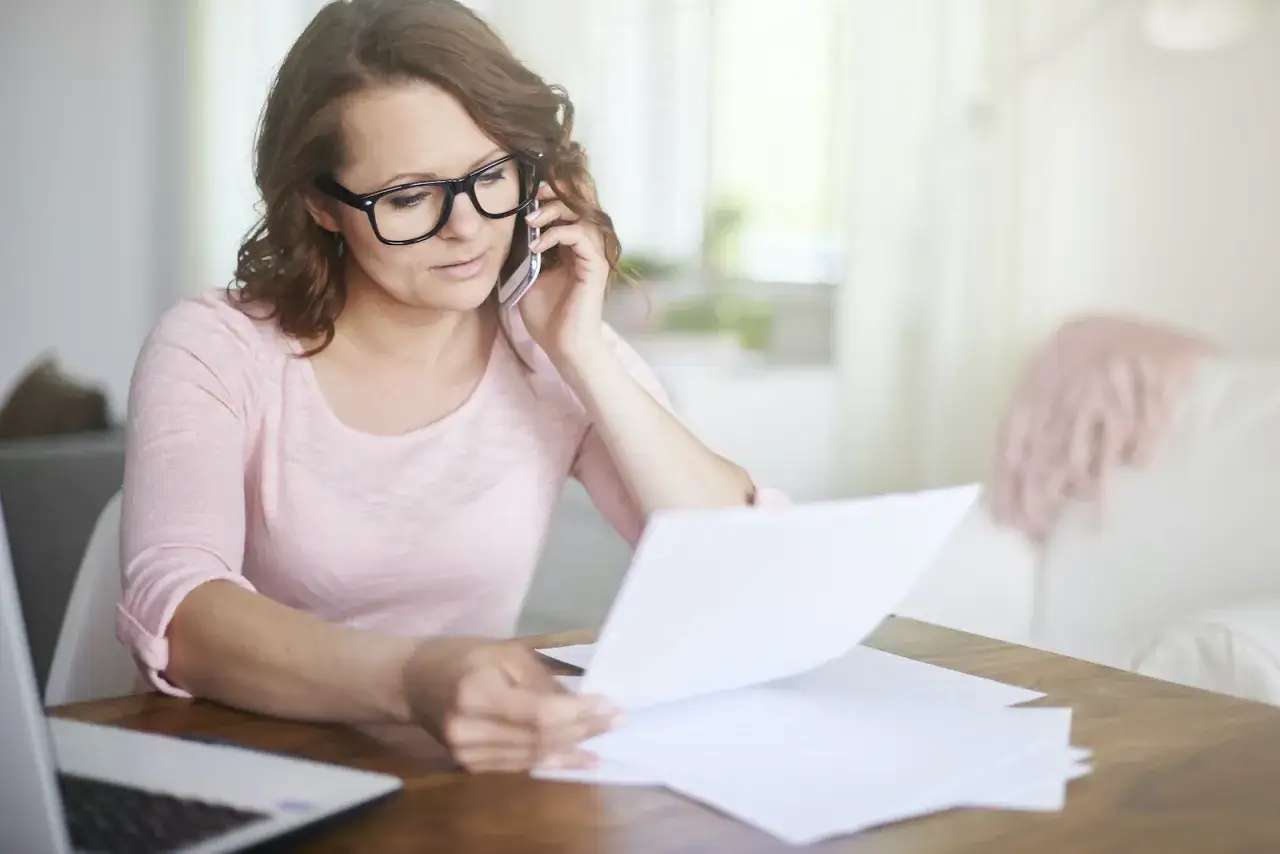Kobieta w okularach rozmawia przez telefon, przeglądając dokumenty. Prawdopodobnie zajmuje się sprostowaniem świadectwa pracy.