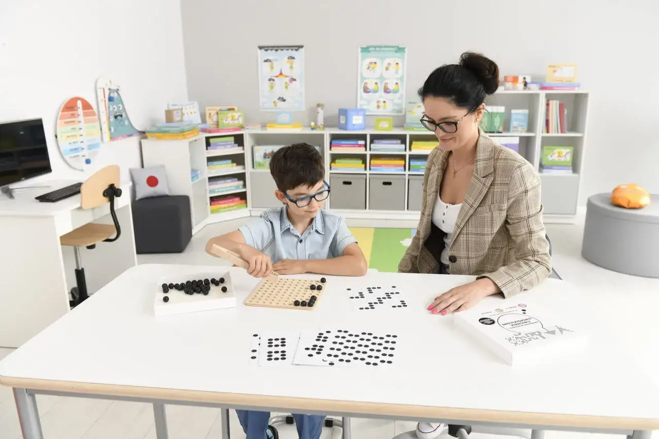 Chłopiec z nauczycielem podczas lekcji w domu. Dziecko układa koraliki na planszy, ćwicząc koordynację wzrokowo-ruchową. Homeschooling w praktyce.