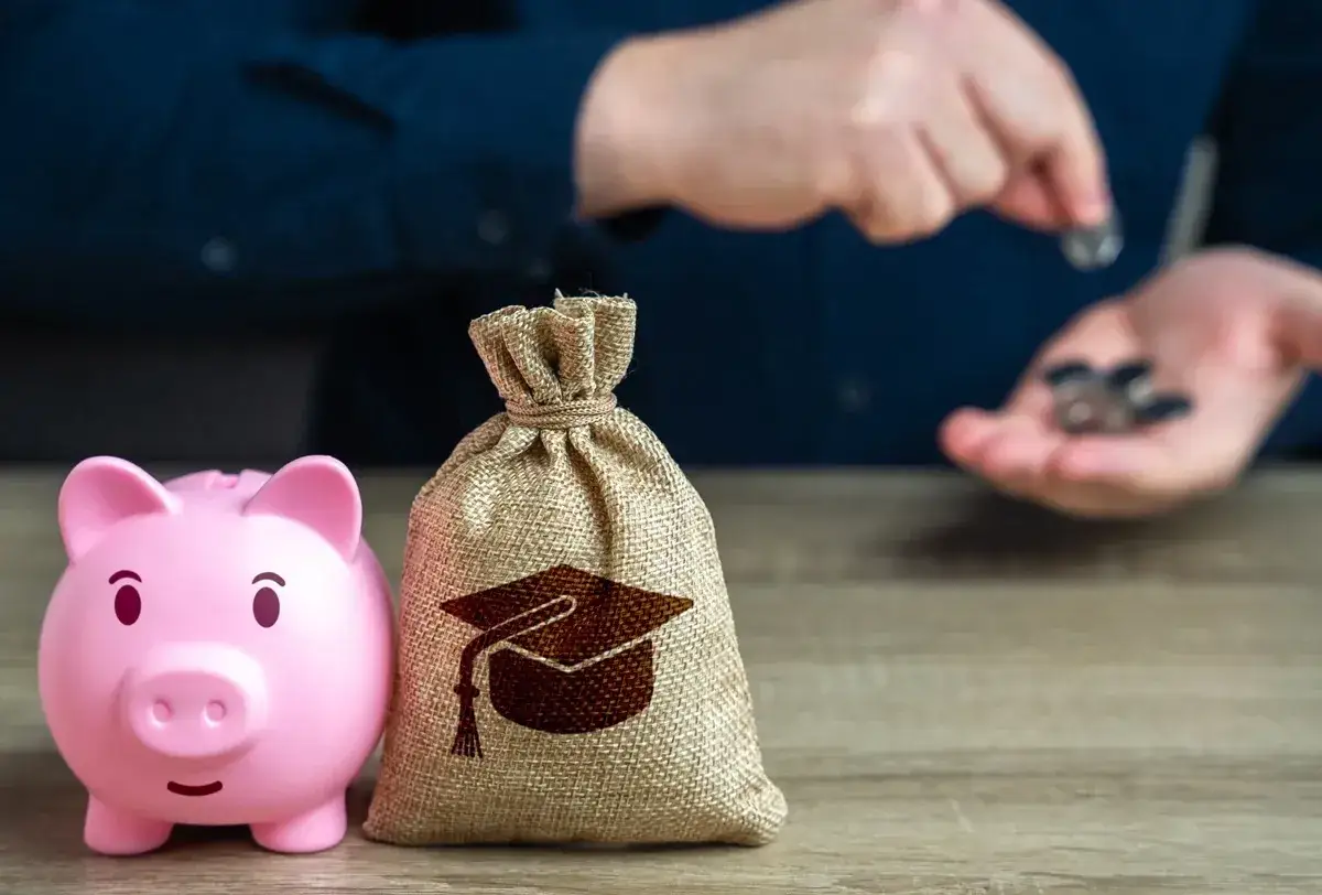 Różowa świnka-skarbonka i worek z symbolem czapki absolwenta. Ręka wrzuca monety, sugerując, jaki dochód do stypendium szkolnego.