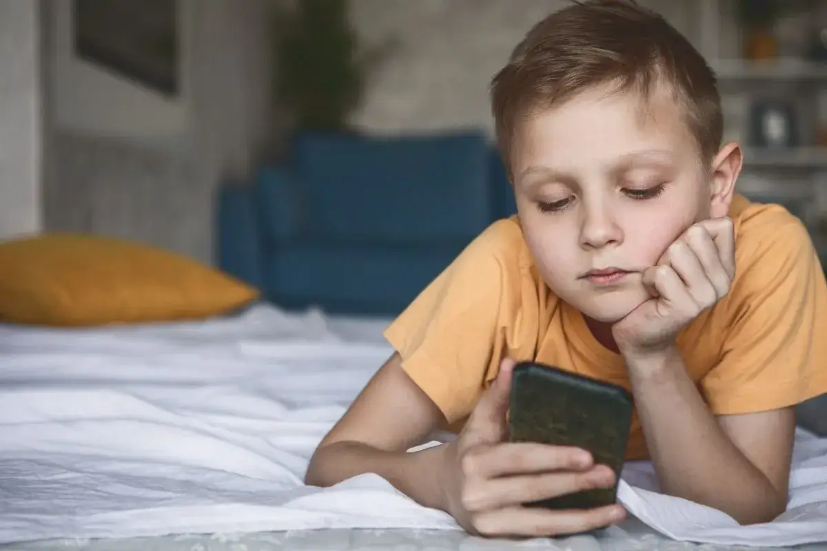 Chłopiec leży na łóżku, trzymając telefon. Jego wzrok jest skupiony na ekranie, a mina sugeruje zamyślenie, być może związane z sekstingiem.