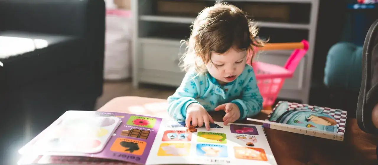 Dziewczynka z niepełnosprawnością intelektualną w stopniu umiarkowanym z zainteresowaniem przegląda kolorową książeczkę edukacyjną, wskazując paluszkiem na obrazki.