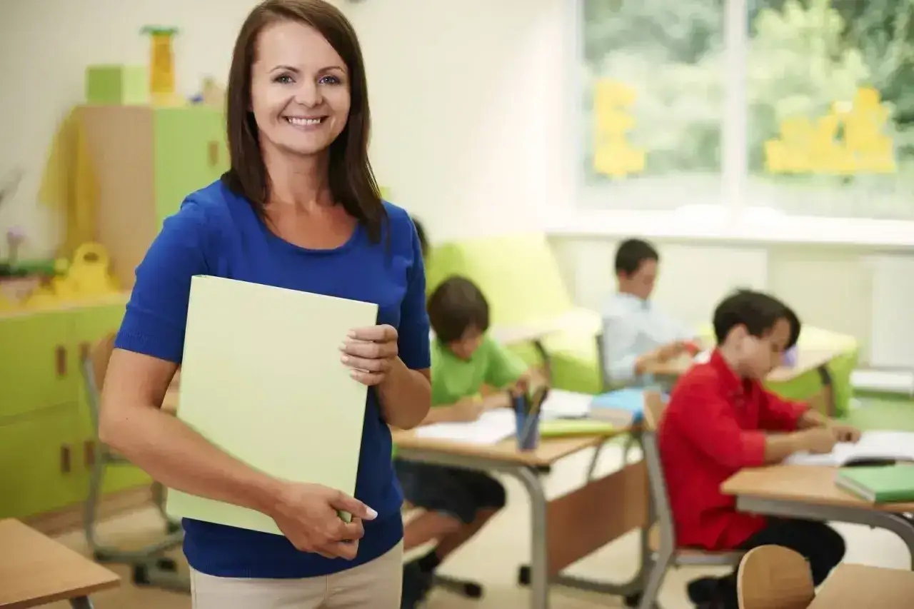 Uśmiechnięta nauczycielka z teczką w ręku, w tle dzieci pracujące przy ławkach. To obrazek z życia szkoły, gdzie pedagogika odgrywa kluczową rolę.