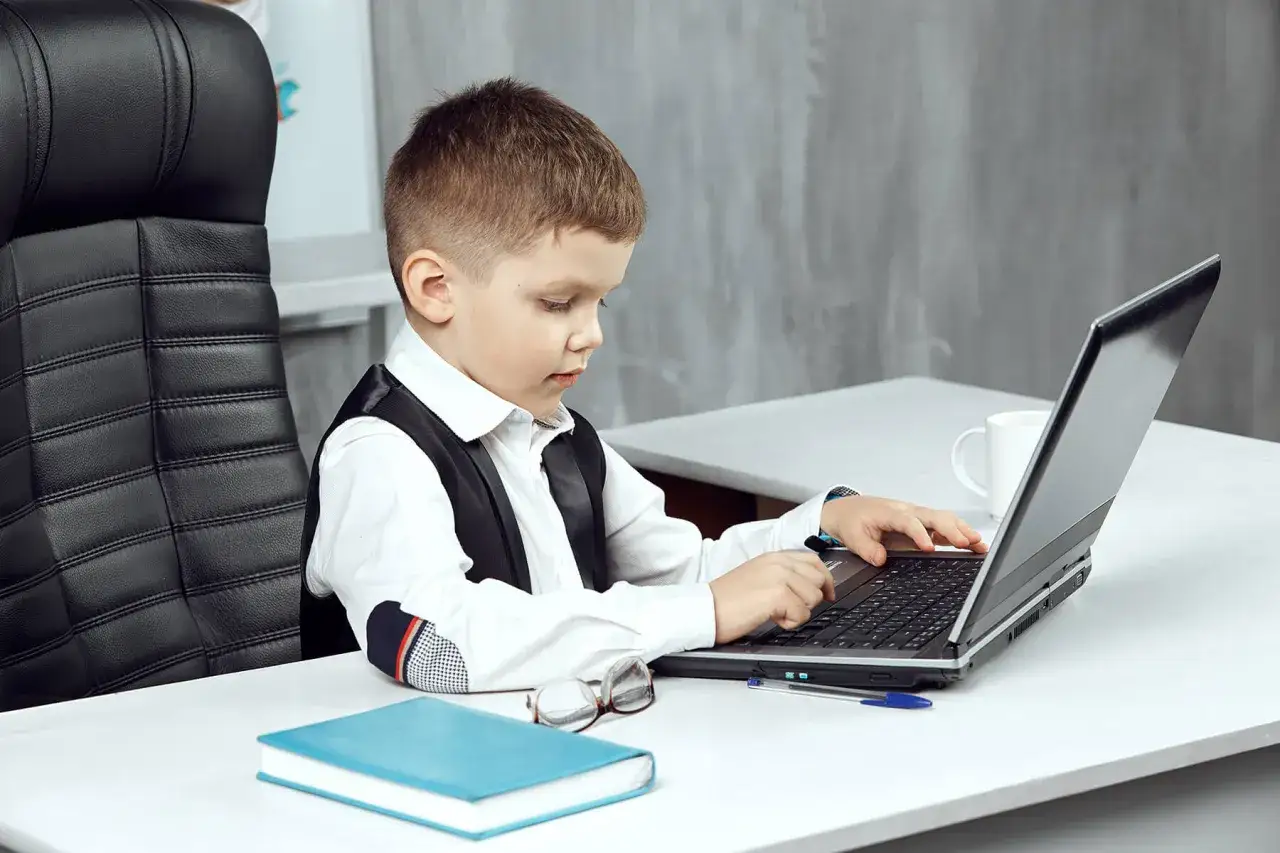 Chłopiec z autyzmem, ubrany w elegancką kamizelkę, skupiony na pracy przy laptopie. Obok leży książka i okulary.