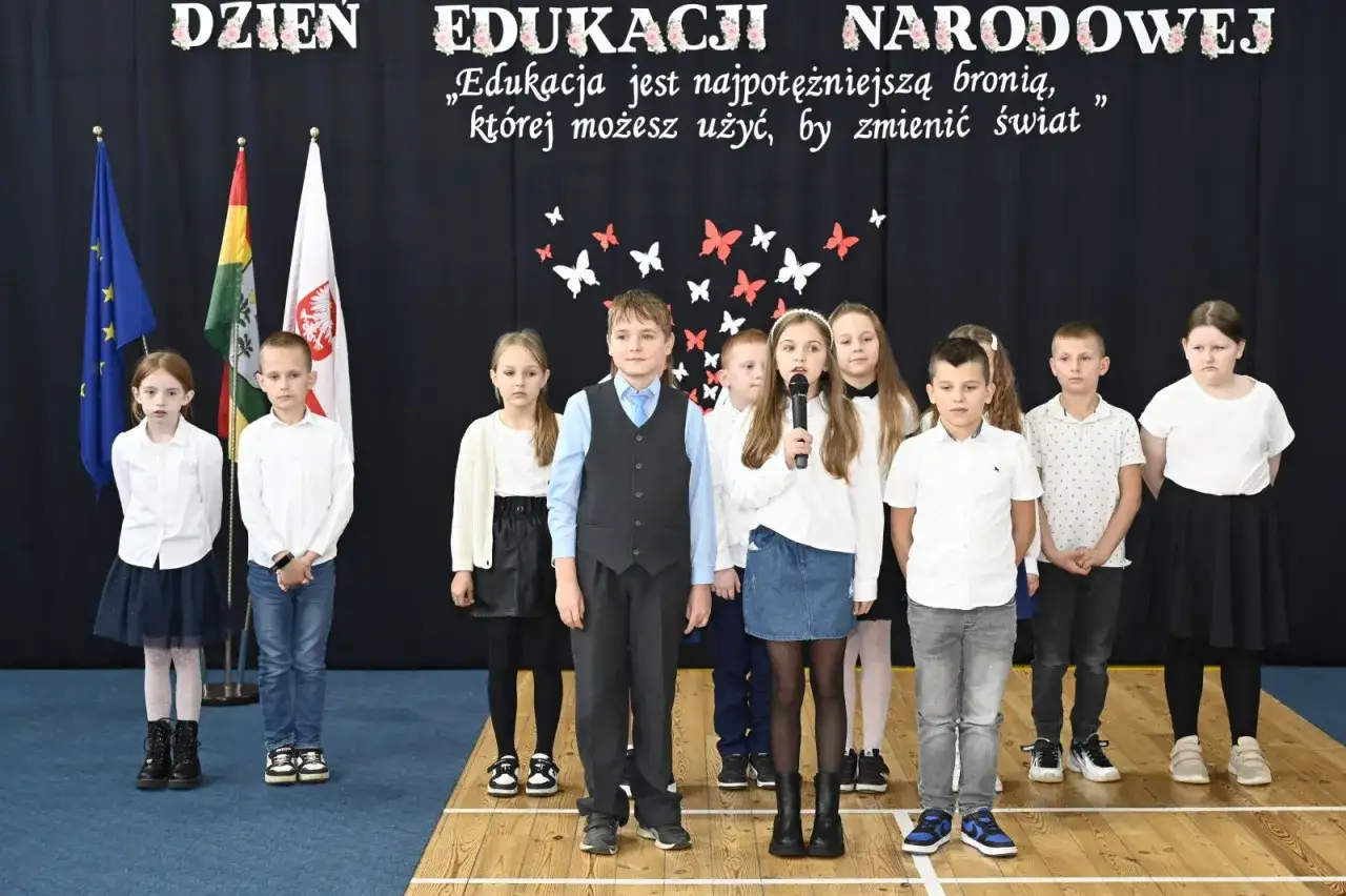 Dzieci na scenie podczas uroczystości z okazji Dnia Edukacji Narodowej. Dziewczynka z mikrofonem przemawia.