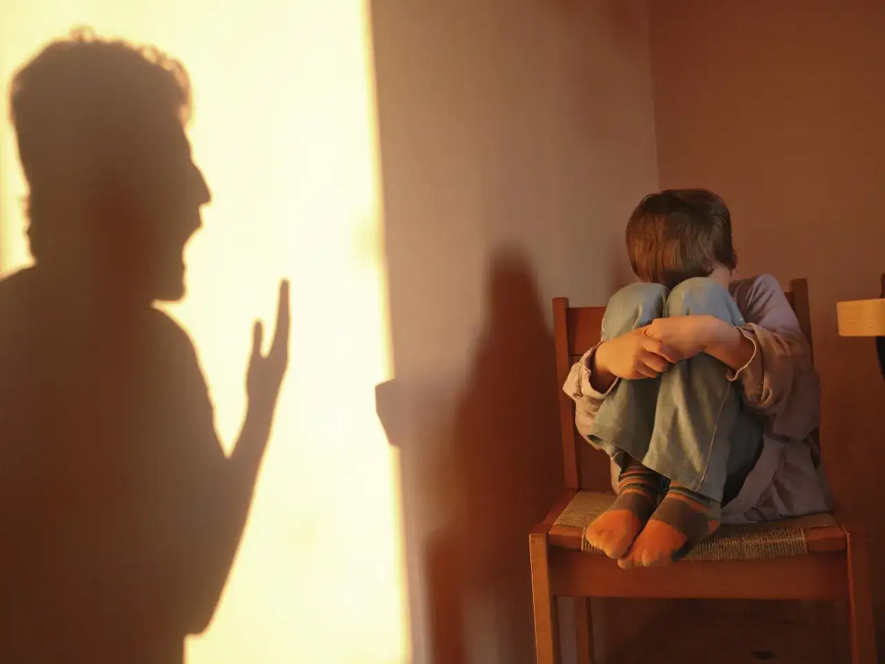 Cień krzyczącej postaci rzuca się na ścianę, podczas gdy dziecko skulone na krześle próbuje się ukryć. To obraz tego, czym jest bullying.