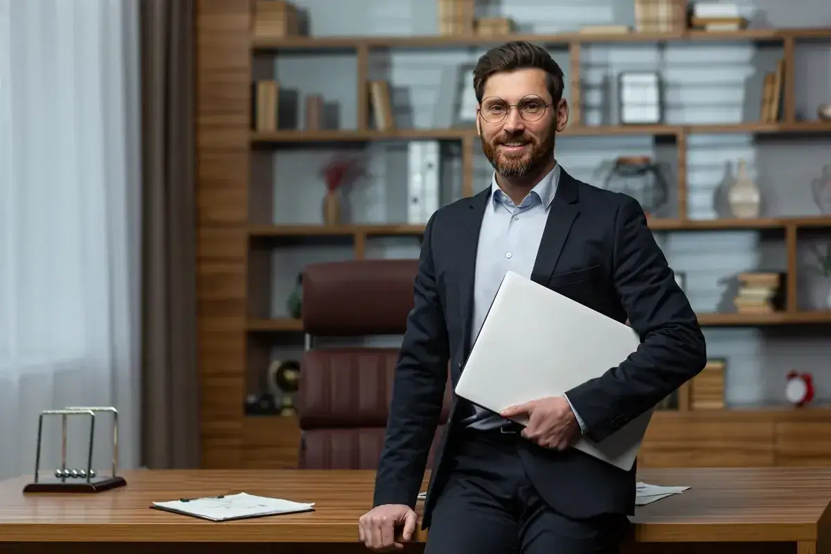 Uśmiechnięty dyrektor szkoły w garniturze, trzymający laptopa. Zastanawia się, ile trwa kadencja dyrektora szkoły.