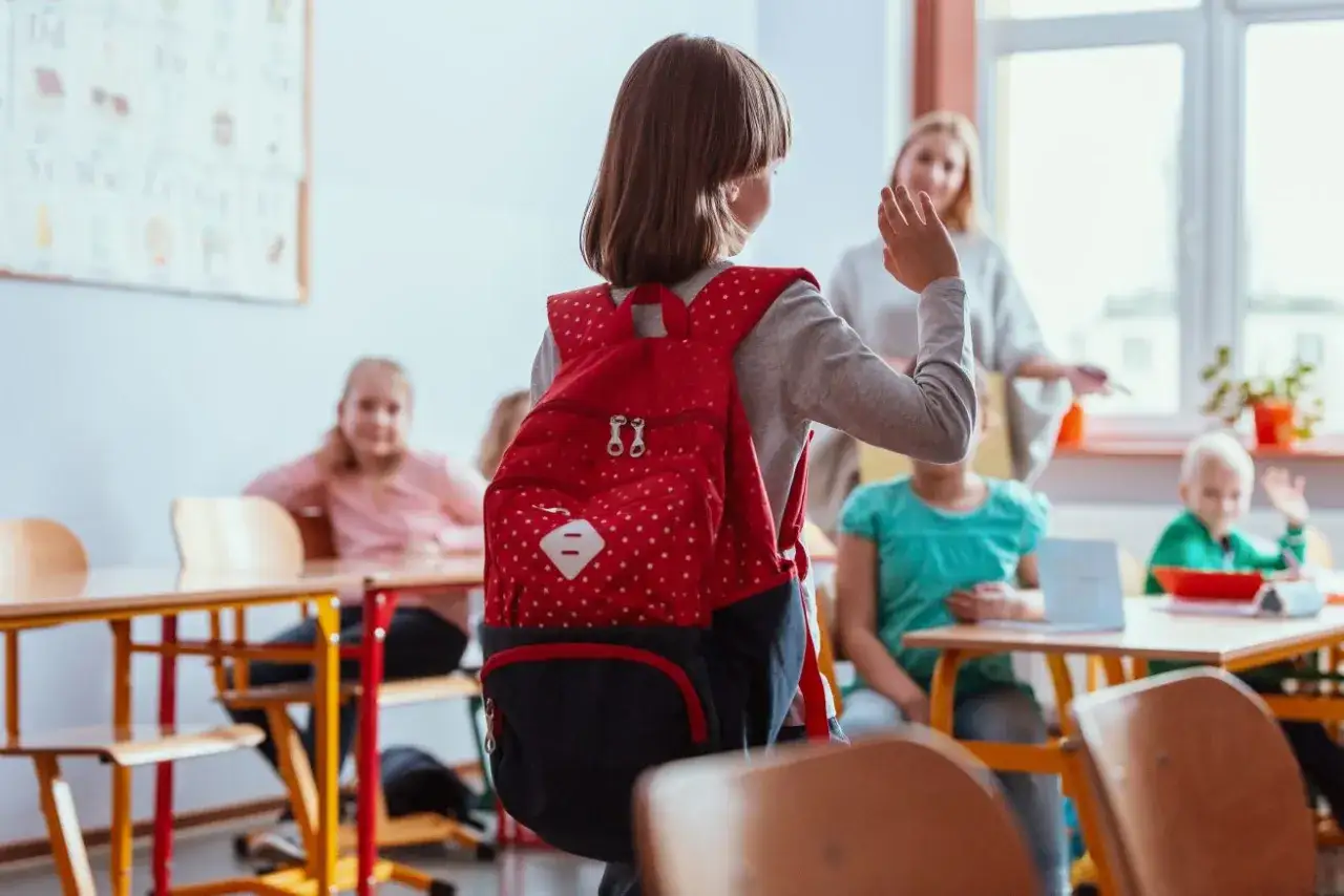 Dziewczynka z plecakiem waha się przed wejściem do klasy, czując fobię szkolną. Nauczycielka i inni uczniowie czekają.