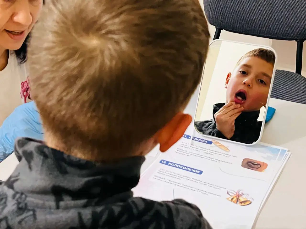 Chłopiec z niepełnosprawnością intelektualną w stopniu lekkim ćwiczy artykulację przed lustrem, pod okiem terapeuty.