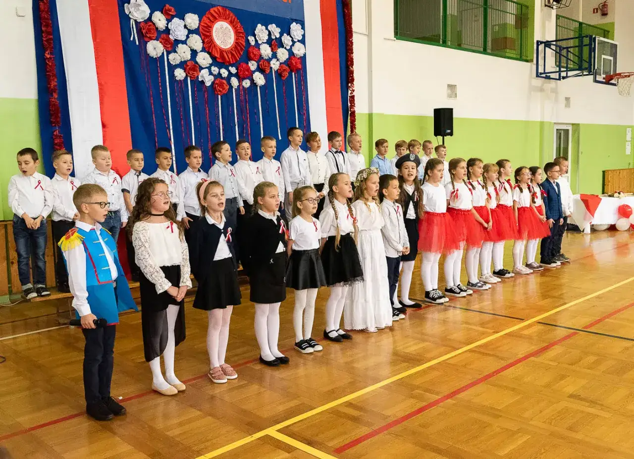 Dzieci w strojach galowych, z biało-czerwonymi akcentami, śpiewają na tle flagi. Widać dumę i patriotyzm w ich oczach.