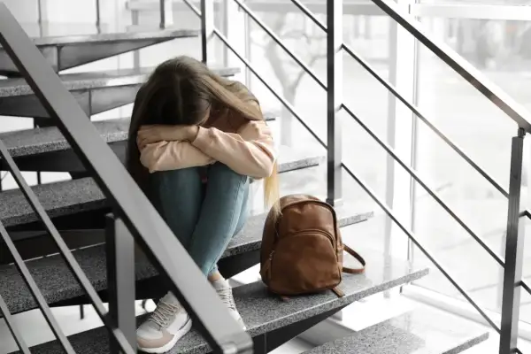Smutna dziewczyna siedzi na schodach, przytulając kolana. Obok niej leży plecak. Martwi się o warunkowe przejście do następnej klasy.