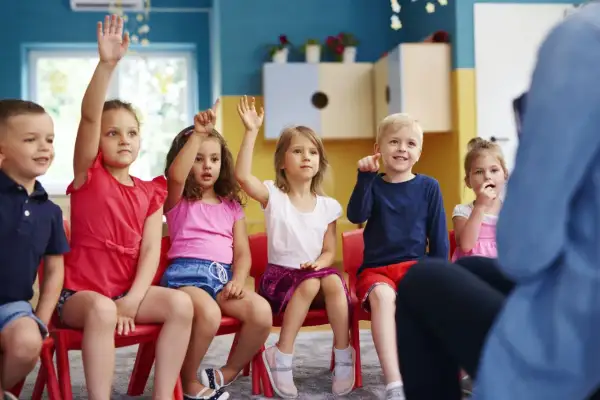 Dzieci na czerwonych krzesłach podnoszą ręce, jakby odpowiadały na pytanie, czy zerówka jest obowiązkowa.