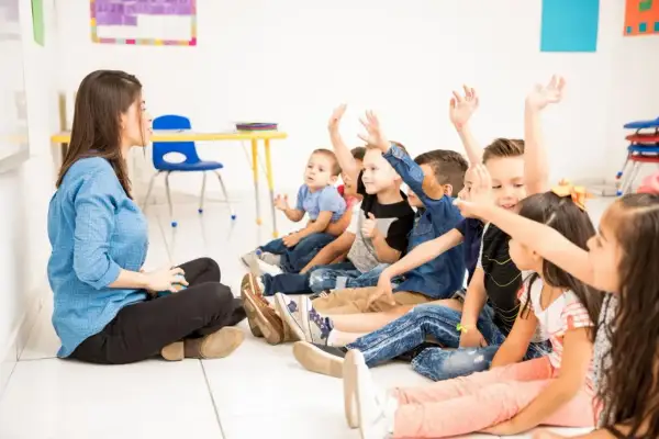 Nauczycielka siedzi na podłodze, a dzieci z podniesionymi rękami odpowiadają na pytania. Widać zaangażowanie i wysoką frekwencję w szkole.