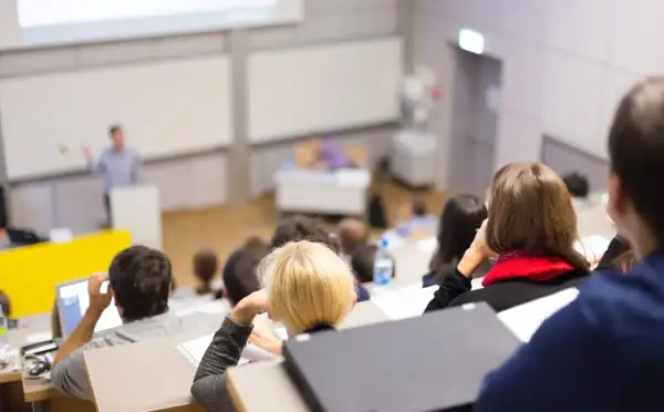 Nauczyciel akademicki prowadzi wykład w auli, studenci słuchają, niektórzy notują.