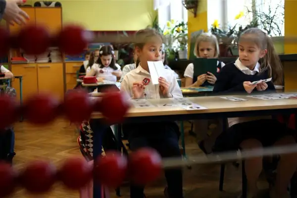 Dzieci siedzą w ławkach, ucząc się, kiedy idą do szkoły. Jedna dziewczynka trzyma kartę z cyfrą.