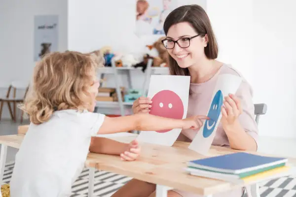Uśmiechnięta psycholog z okularami pokazuje dziecku karty z buźkami. To część przygotowania pedagogicznego dla psychologów.
