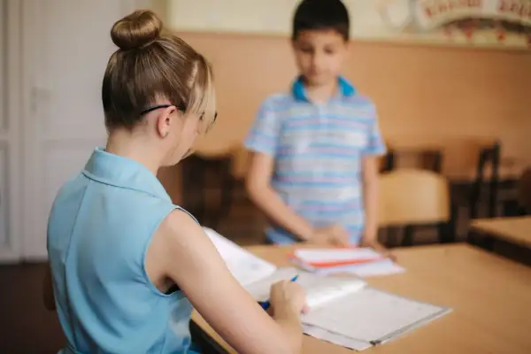 Nauczycielka z kucykiem przygląda się zeszytom, podczas gdy uczeń stoi w tle. Czy to zapowiedź podwyżki dla nauczycieli? Najnowsze wiadomości wkrótce.