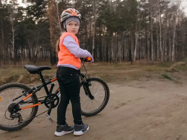 Dziewczynka w kasku i kamizelce przygotowuje się do egzaminu na kartę rowerową, stojąc obok swojego roweru na leśnej ścieżce.