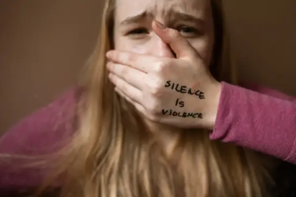Przerażona dziewczyna zakrywa usta dłonią z napisem "Silence is violence". To obraz patologii społecznej, która odbiera głos.