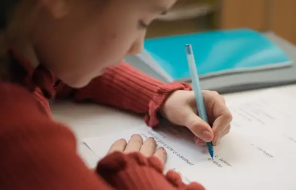 Dziecko w czerwonym swetrze pisze długopisem po karcie pracy, starając się o dopuszczający ocena.