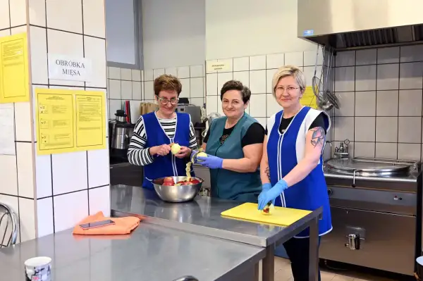 Trzy kobiety w fartuchach obierają jabłka w kuchni. Jedna z nich, intendentka, przygotowuje owoce do dalszej obróbki.