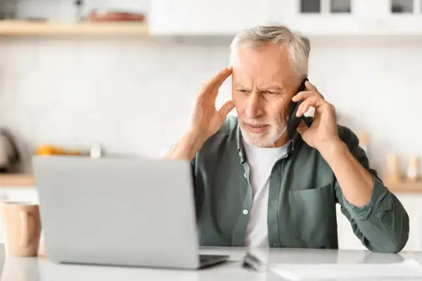 Zmartwiony mężczyzna rozmawia przez telefon, trzymając się za głowę. Obok laptopa i kubka. Wygląda na to, że jego urlop z powodu siły wyższej został zakłócony.